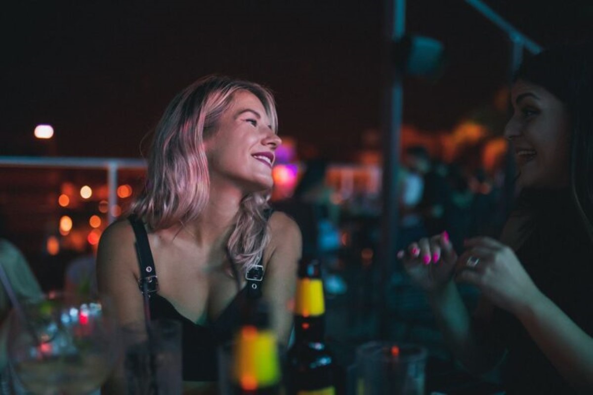 Two women enjoying an evening at Memories rooftop bar in Larnaca; one with pink-tinted hair looking up and the other talking animatedly, surrounded by dim lighting and bar essentials